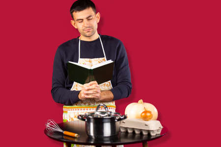 Young man in the kitchen, reading a cookbook of recipes. Gender stereotypes. Photo on a pink background with side space.の写真素材