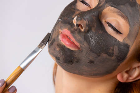Enlarged photo of a young woman with closed eyes applying a clay mask to her face with a brush. Photo on white backgroundの写真素材