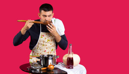 Tasting soup. Handsome young man tasting soup from a saucepan. Photo on a pink background with empty side space. Gender stereotypes conceptの写真素材