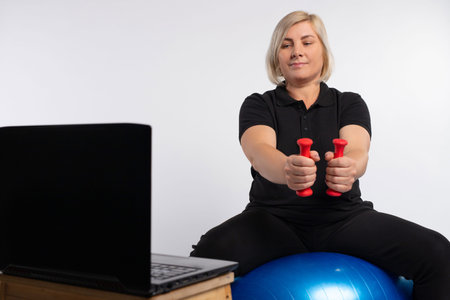 Senior woman doing fitness exercises with a virtual classroom sitting on a fitness sword holding...