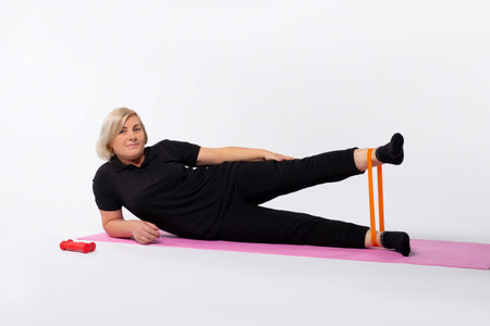 Athletic senior woman doing fitness workout at home with resistance rubber band lying on mat....