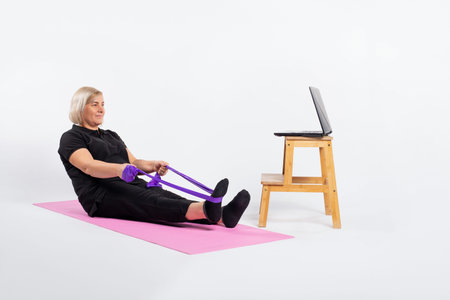 Senior woman sitting on a fitness mat in front of the computer and doing exercises with an elastic...