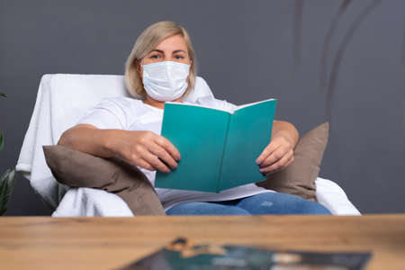 Senior woman in a medical mask is resting at home in a comfortable chair with her favorite book. Pandemic and quarantine concept.の写真素材