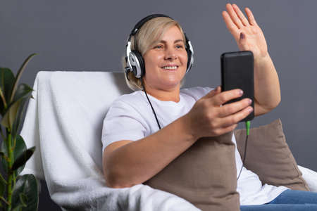 Senior woman in headphones talking on a video call on the phone while sitting in a comfortable chair. Senior online concept and pandemic concept.の写真素材