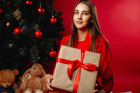 A beautiful girl with fluffy hair next to a Christmas tree and holding a gift. Cute posing for the camera. New Year holidays concept on red background.の写真素材
