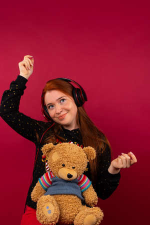 A cute girl with a bear, listens to music in big headphones and dances. Photo on a pink background. Spending time concept.の写真素材