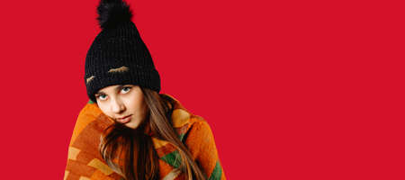Banner, long format. Portrait of a cute girl in a winter hat and wrapped in a warm plaid, posing on a red photo with side space. Winter conceptの写真素材