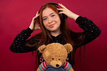 Red-haired girl with a bear. Listens to music while holding big headphones, posing and smiling. Photo on a pink background.の写真素材