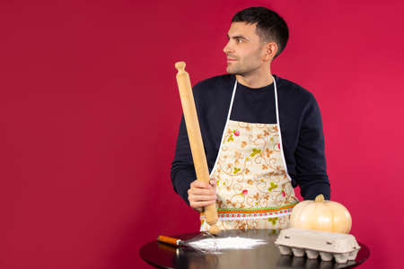 The young man looks thoughtfully to the side with a rolling pin in his hands. Photo on a pink background with empty side space. The concept of gender stereotypes.の写真素材