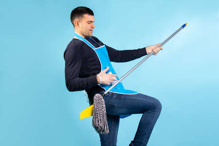 Cheerful man in an apron, having fun and dancing with a mop while cleaning the house. Gender stereotypes.の写真素材