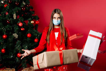 A girl in a mask, in pajamas by the tree, a bunch of gifts in the air. Girls power, superhero. Holidays pandemic concept. Photo on a red background.の写真素材