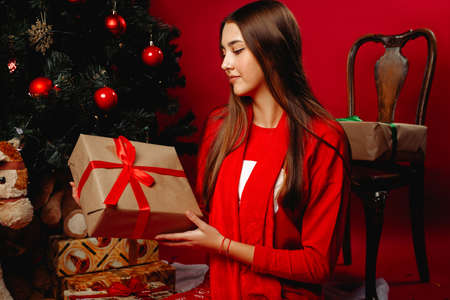 A cute girl at the Christmas tree, holding a gift box in her hands. The girl looks at the gift with interest. New Year holidays concept on red background.の写真素材