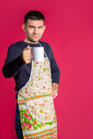 Young man with a cup of tea and in a kitchen apron on a pink background. The concept of gender stereotypes.の写真素材