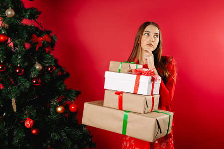 A girl with at home by the tree holding gift boxes. Concept of New Years gift, winter shopping. Red background.の写真素材