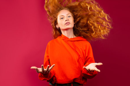 A red-haired girl with fluttering curly hair and arms to the sides. Looks at the camera on a red background. High quality photoの写真素材