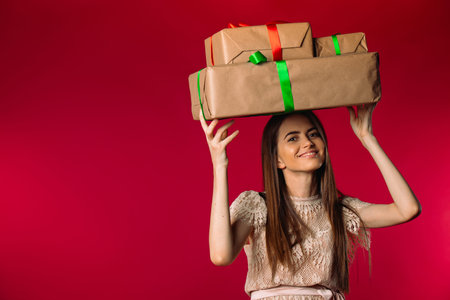 A beautiful young girl with long hair holds large gift boxes on her head and smiles. Photo on a pink...