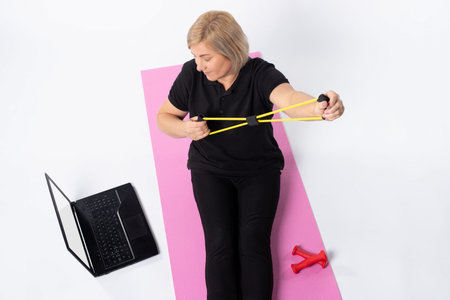 Photo from above. Senior woman holds a resistance band on the carpet in front of a laptop. High...
