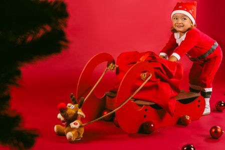 A little girl in New Year's outfit happily pushes a sled and a bag of gifts. Red background. Christmas. High quality photoの写真素材
