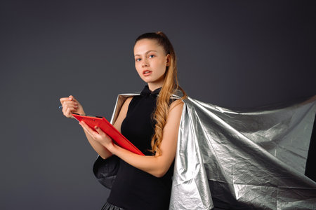 Serious lady, superhero in a gray cloak , holding documents in red folder on a gray background. High quality photoの写真素材