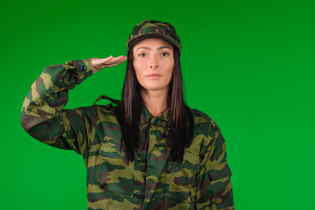 Portrait of a woman in military uniform. Holds hand to temple. Military greeting. Green background. High quality photoの写真素材