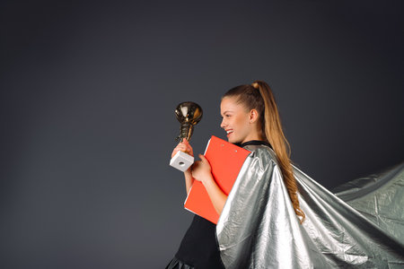 Superheroine, businesswoman with a red folder and a champions cup in hands being satisfied of trophy on a gray background. High quality photoの写真素材
