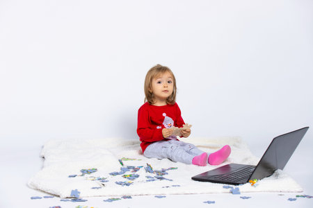 Little girl plays with puzzles sitting on white blanket with a laptop, looking at the camera on a...