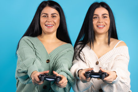 Two twin sisters with joysticks on a blue background posing, smiling and looking at the camera. High quality photoの写真素材