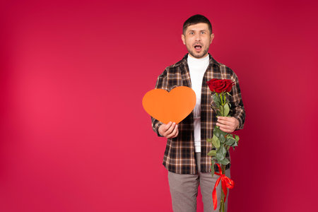 Surprised man with St. Valentine's Day greeting card in form of heart and red roses on a red background with empty side space. High quality photoの写真素材