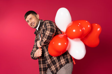 A guy wrinkles his face carrying a bunch of red heart-shaped balloons on red background. St.Valentine's holiday concept. High quality photoの写真素材