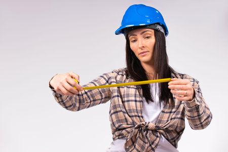 A young woman in a blue hard hat looks at a tape measure in her hands. White background.