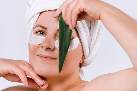 Portrait of caucasian elderly woman with fair skin, cosmetic eye patches and aloe vera leaf in front of face, closing an eye. White background.の写真素材