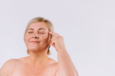 A satisfied woman applies a revitalizing cream under the eyes with her index finger. White background with empty space.の写真素材