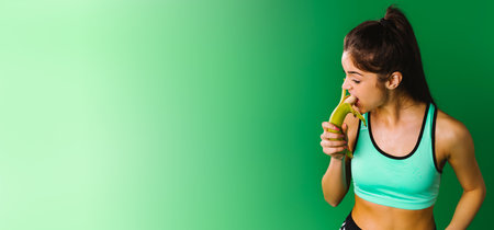 Banner,long format. Healthy food concept. Fitness girl in sportswear on a green background eats a banana for strength and energy. Healthy food concept.の写真素材
