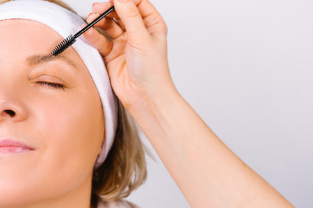 Enlarged photo where the hand of the master makes the lamination of the eyebrows of a woman with a bandage on her head. Fashion trends on a white background.の写真素材