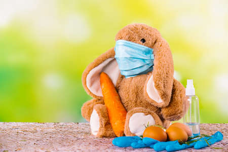 Rabbit on green background with mask for coronavirus, eggs, big carrot, protective gloves and disinfectant around the composition. Happy easter and stay home.の写真素材