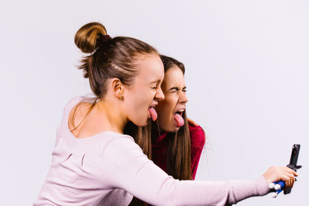 Two girlfriends fool around in live streaming with a smartphone on selfie stick show their tongues....