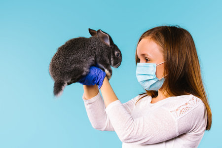 Blonde girl in a disposable medical mask and blue latex gloves holds a fluffy rabbit in her arms,...