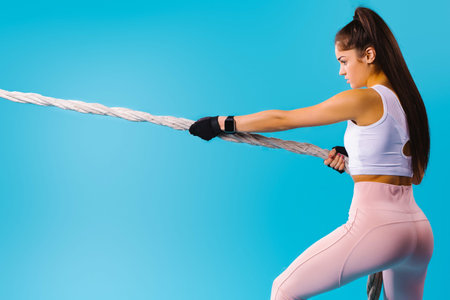 Tug of war. Strong sportswoman pulls the battle rope on a blue background with blank space. Powerful...