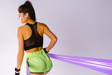 Photo from behind. A girl in fitness shorts and a top trains body mice with an resistance elastic...