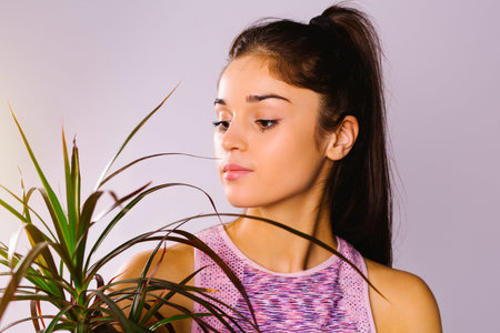 Portrait of a young attractive athletic girl looking at a green indoor flower on a white studio...
