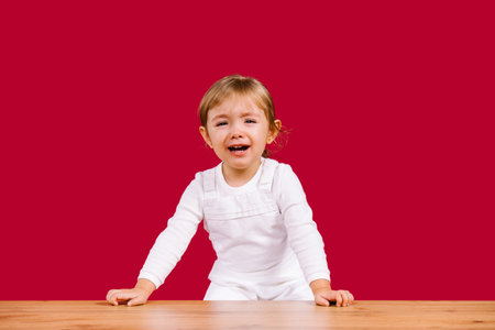 Blonde toddler girl wearing casual white clothes is scared and crying girl standing over wooden table on red backgroundの写真素材