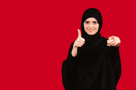 Smiling young arabian muslim woman in hijab black clothes pointing index finger to camera and thumb up on isolated red background. People religious lifestyle conceptの写真素材