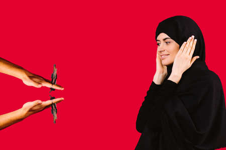 A woman in a black hijab stands over red background while next to a man gives her the keys to a new apartment. Joyful arab woman rejoices at a gift from a man. Red background with empty spaceの写真素材