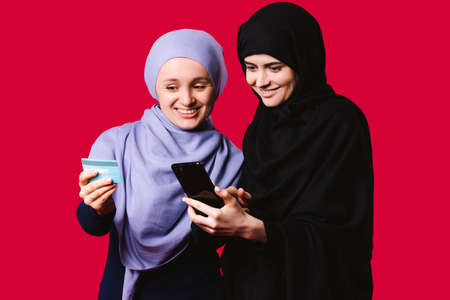 Elegant arab women holding mobile phone and banking card. Businesswomen with stylish hijab paying with credit card while shopping online using mobile phone on red backgroundの写真素材