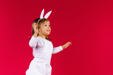 Caucasian liitle blonde girl with white bunny ears jumping on red background, looking up on empty...