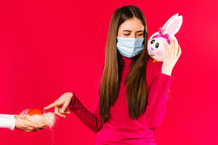 A girl in a medical mask holds a decorative bunny on her shoulder and points with her index finger...