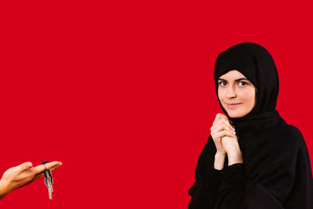 Surprised young girl in black hijab looks at the camera. A mans hand holds out the keys to the apartment. Gift for Muslim women on a red background.の写真素材