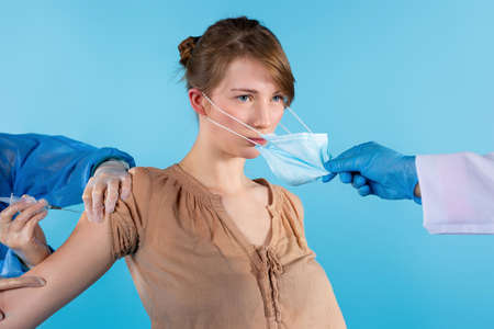 Victory over coronavirus. Pandemic Covid-19. Serious blonde woman after coronavirus vaccine. Doctor hand takes off protective sterile medical mask from face. Happy end on blue backgroundの写真素材