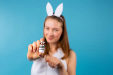 Coronavirus vaccination. Caucasian blonde girl wearing bunny ears in background, holding focused ampoule of covid 19 vaccine in front of camera. Happy Easter, safe, healthy. Pascal pandemic on blueの写真素材
