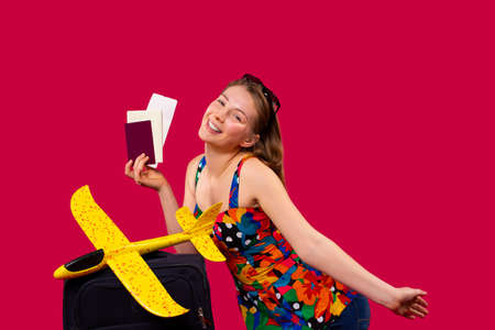 A blonde woman in bright clothes with documents, a suitcase and a bright yellow toy plane, happy to travel and posing on a red background. High quality photoの写真素材